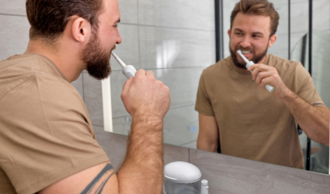 Man with electric toothbrush brushes teeth at 45-degree angle in front of a mirror.